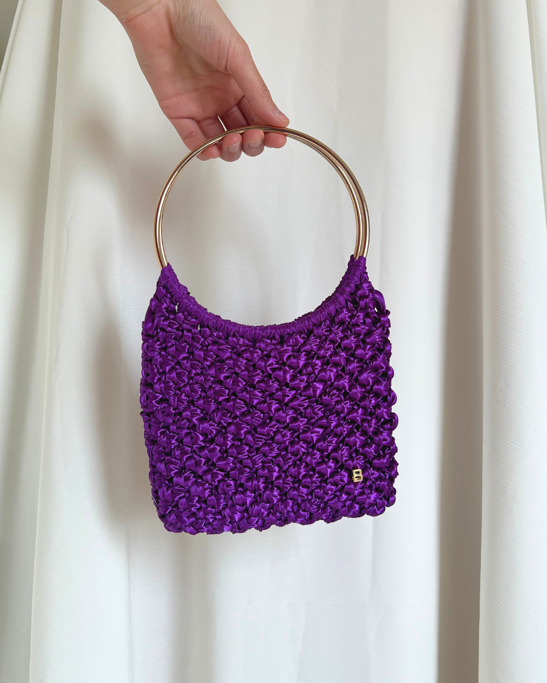 Roma Bag de raso morado con asas doradas brillantes, vista frontal, con la B de Bimbú en la esquina inferior derecha.